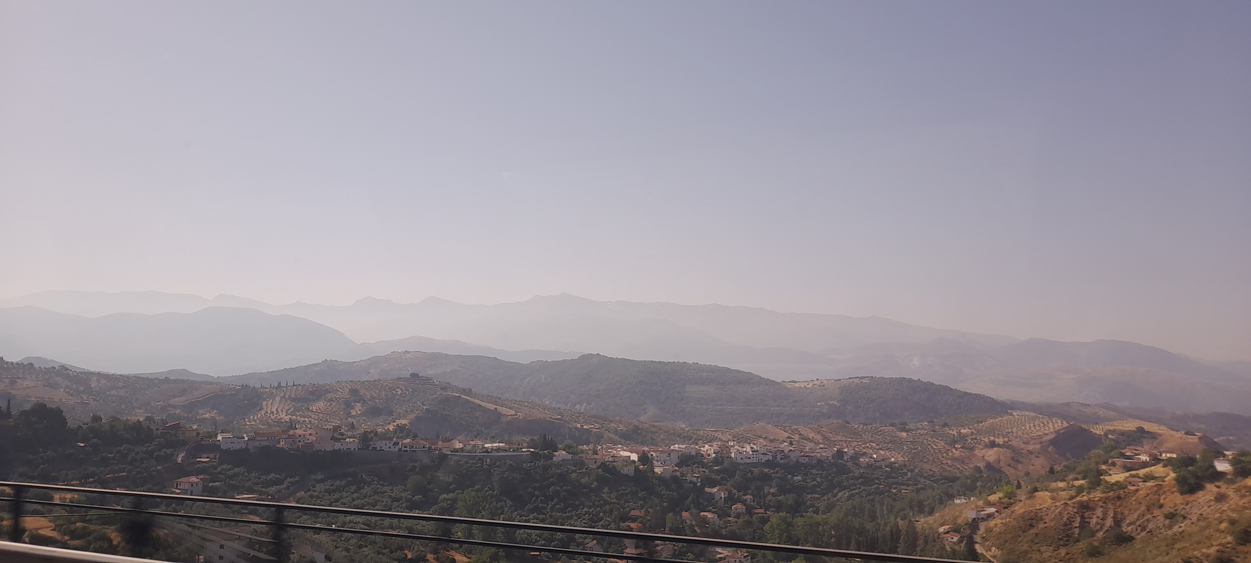 A wide shot of the Andalusian mountains. The foreground is mostly olive and conifer trees leading up to the first ridge; the receding blue ridgelines occur four times over into the far horizon.