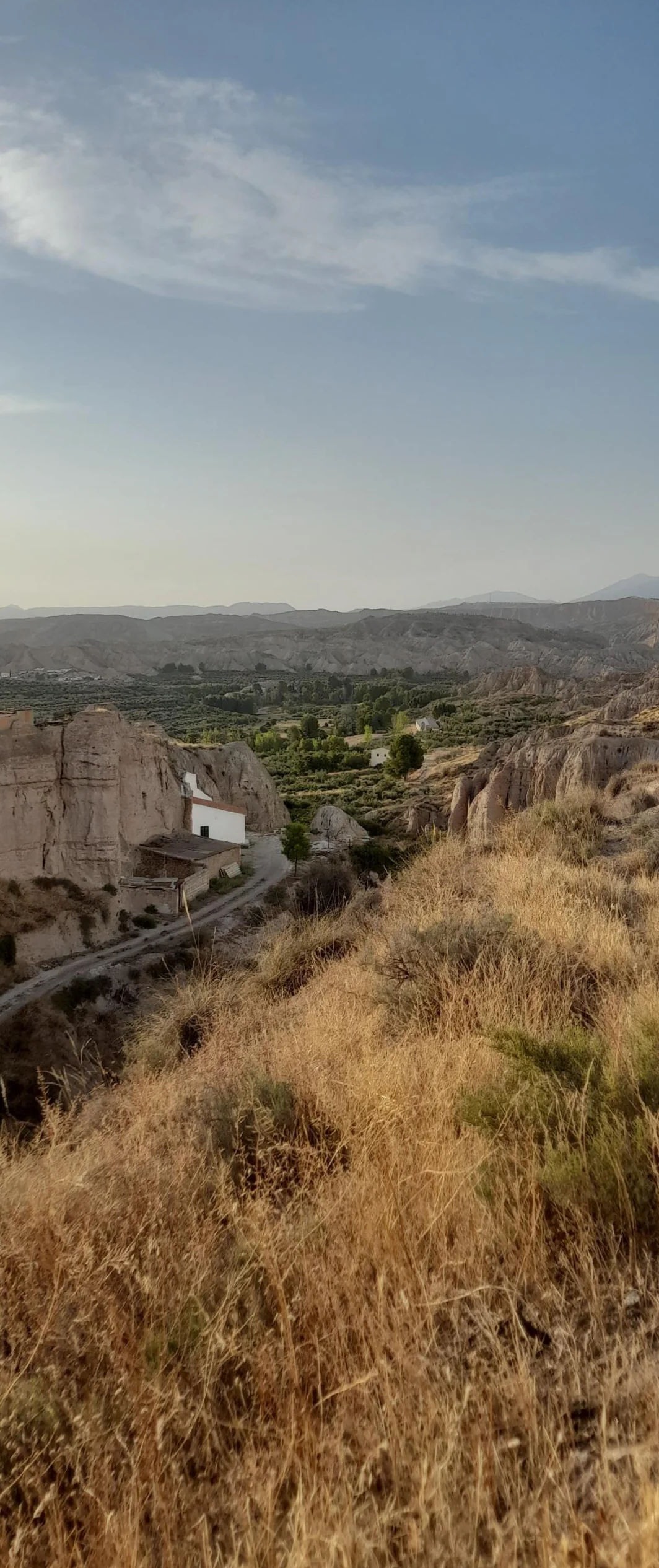 a long shot out from the mirador. dry grass makes up a large portion of the foreground, its a golden straw colour, the sun is to the left. the shot shows a road in the shade of the valley on the left hand side and more sheer cliff face in the soft sedimentary layers on the right. in the distance are three rows of mountain range with the flat center of the landscape filled with various trees and white buildings dotted throughout.