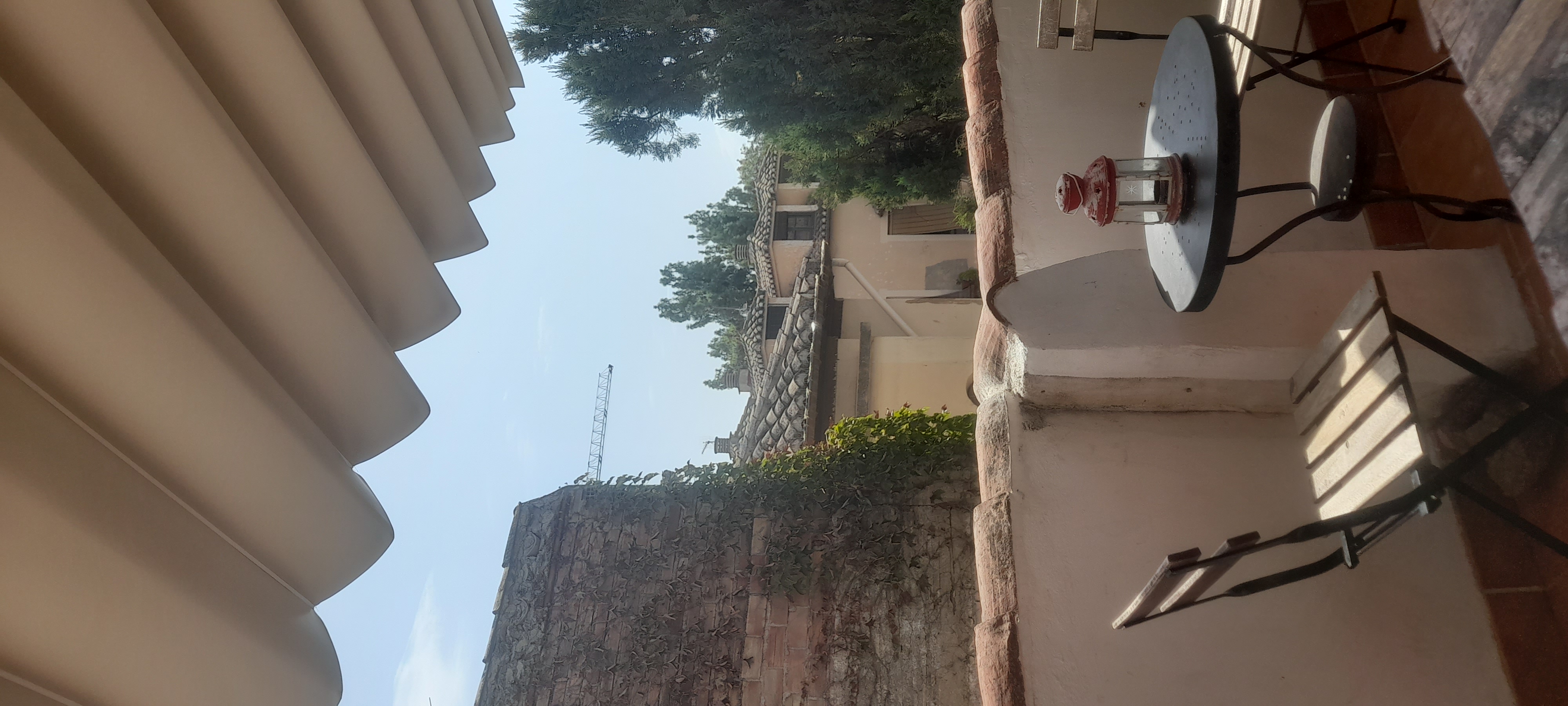 From the hostel balcony, two slim folding chairs with a round table. A red lantern-style lamp is on the table. The balcony wall is a short, white plaster wall topped with clay roof tiles typical of Spain. In the background is a brick wall covered in vines on the left and a conifer on the right. Further into the background are several small dormer roofs with the same clay, half-round tiles; they are backed by more conifers and the arm of a crane poking out. There are no clouds in the stark blue sky. 